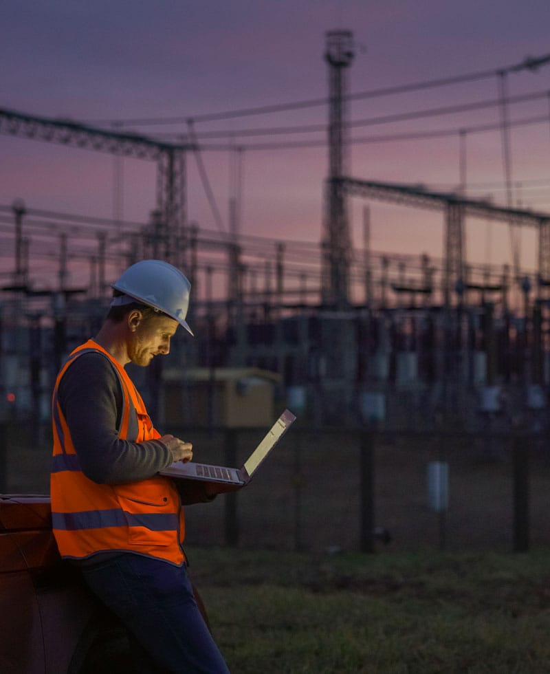Un technicien des services publics, coiffé d'un casque de chantier et vêtu d'un gilet de sécurité, surveille un ordinateur portable dans une sous-station électrique au crépuscule.