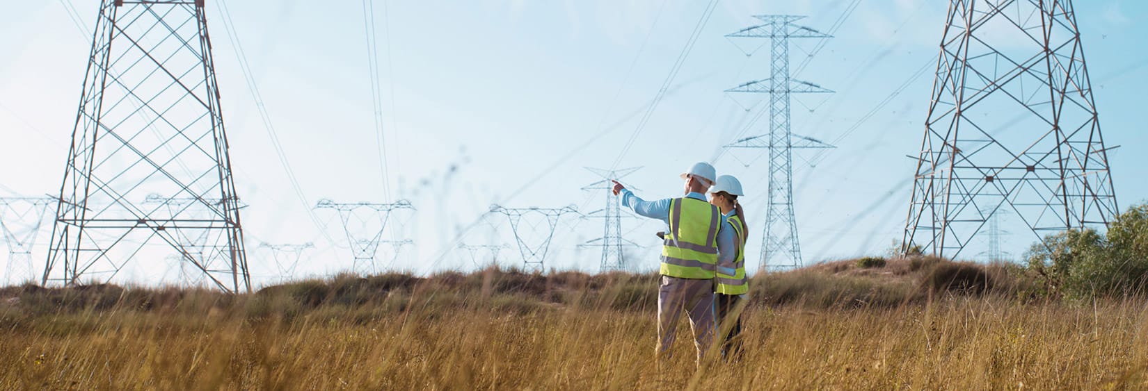 Deux travailleurs des services publics, vêtus de gilets de sécurité et de casques, inspectent des pylônes de transmission d'énergie dans un champ herbeux.